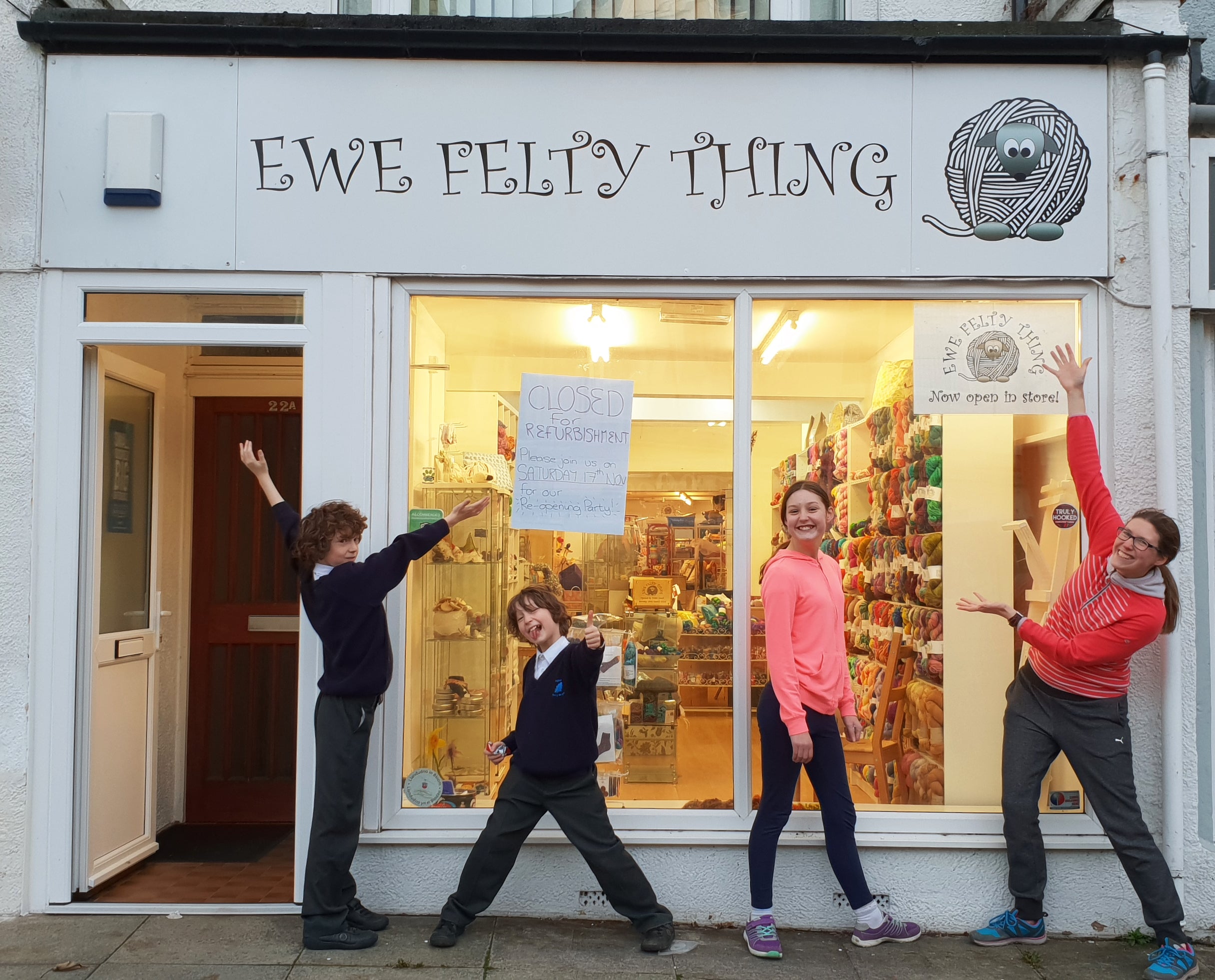 A family stand proudly in front of a yarn shop, pointing at a sign saying Ewe Felty Thing.