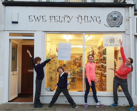A family stand proudly in front of a yarn shop, pointing at a sign saying Ewe Felty Thing. 