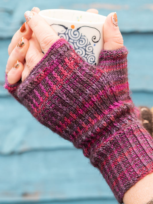 Hands holding a mug, wearing hand-knitted fingerless mitts in a variegated red and purple yarn. 
