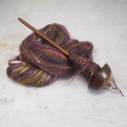 A top whorl drop spindle wrapped with handspun yarn, lying on a bed of purple fibre. 