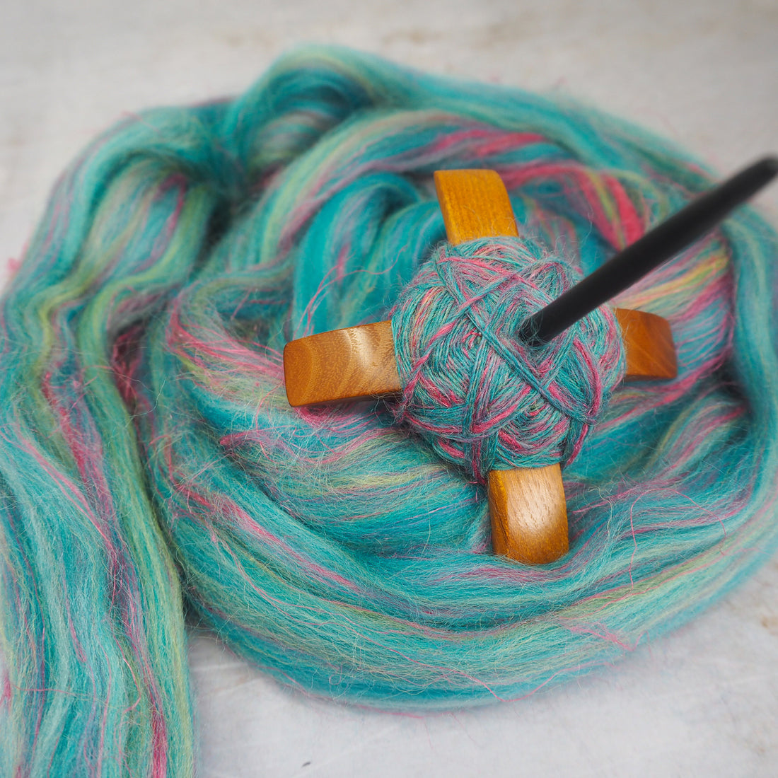 A turkish spindle wrapped with hand-spun yarn, lying on a bed of turquoise fibre. 