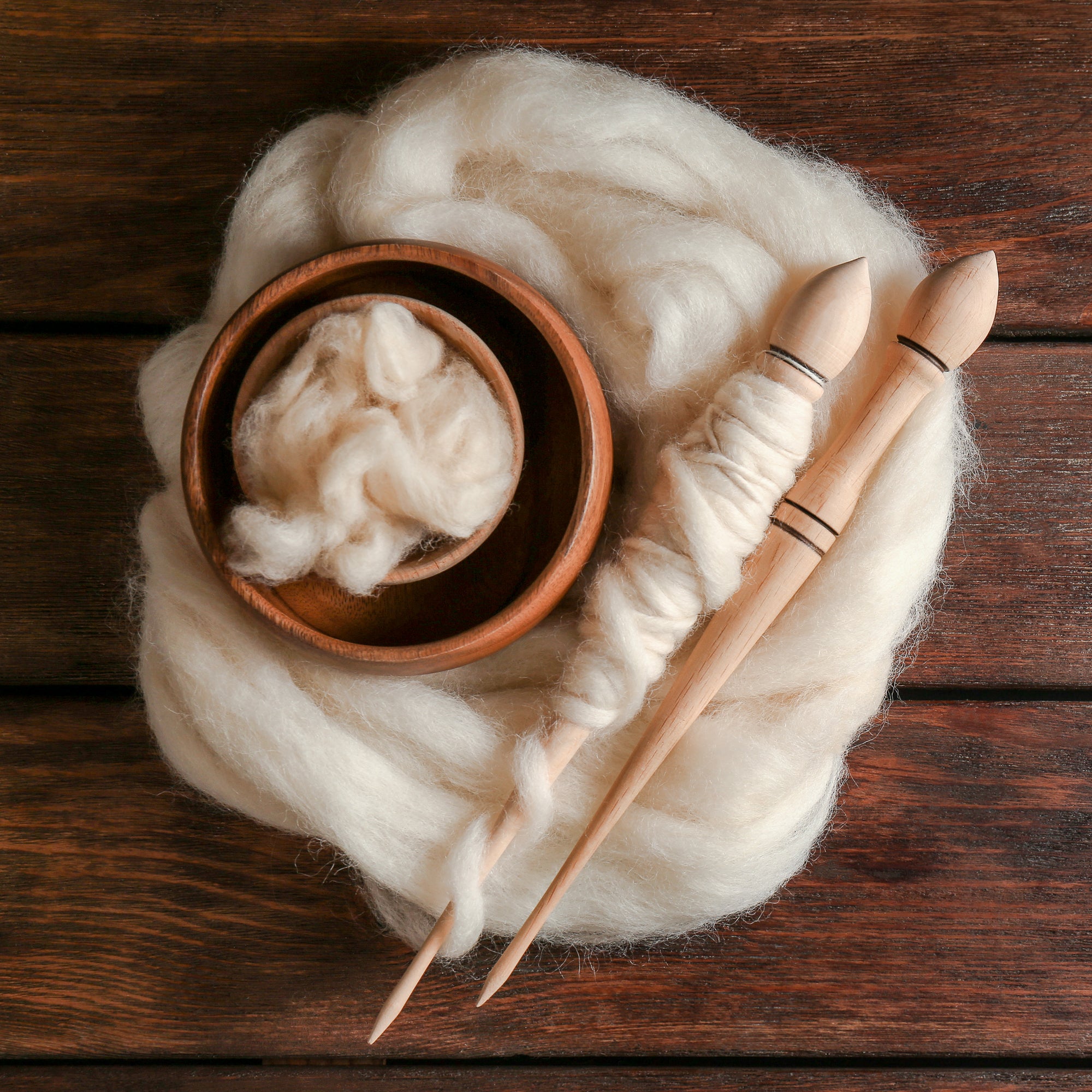 Two Russian spindles lying on a bed of unspun fibre, next to a small wooden bowl. 