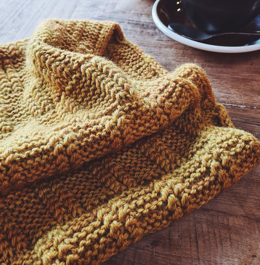 A beautifully textured cowl in soft, golden yarn next to a cup of coffee